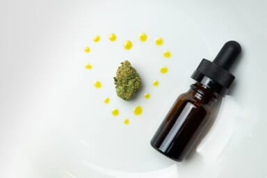 A CBD or THC tincture dropper on a white plate next to a marijuana nug and drops of oil made into a heart surrounding the tincture bottle.